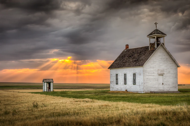 A old church building in a field. Above sunlight peeks through grey clouds.