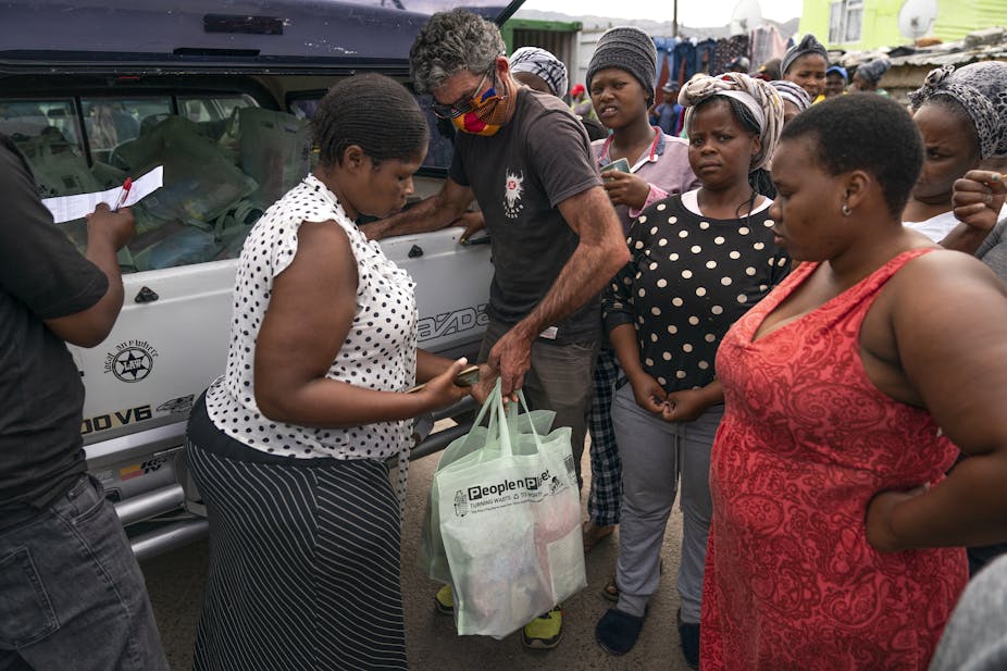 Standing next to a truck, a man wearing a face mask hands a bag to a woman and other people stand nearby, looking on
