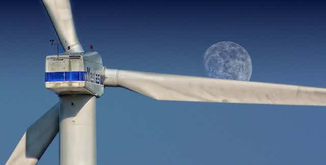 The blades of a wind turbine are seen against a blue sky with a full moon