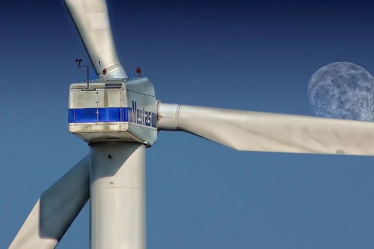 The blades of a wind turbine are seen against a blue sky with a full moon