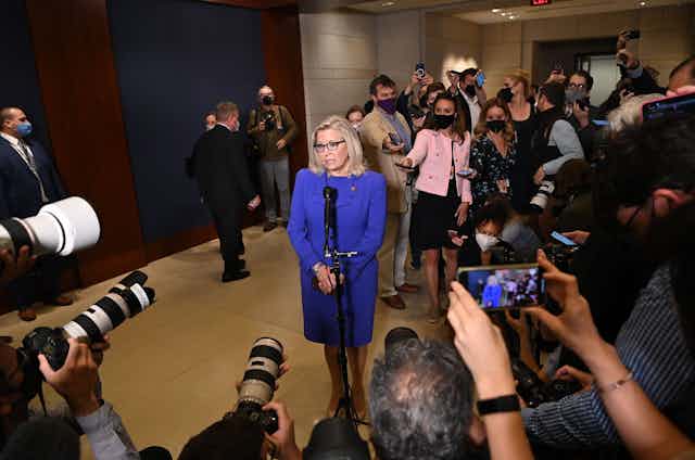 Rep. Liz Cheney, in a bright blue suit, speaking at a microphone while encircled by reporters and photographers.