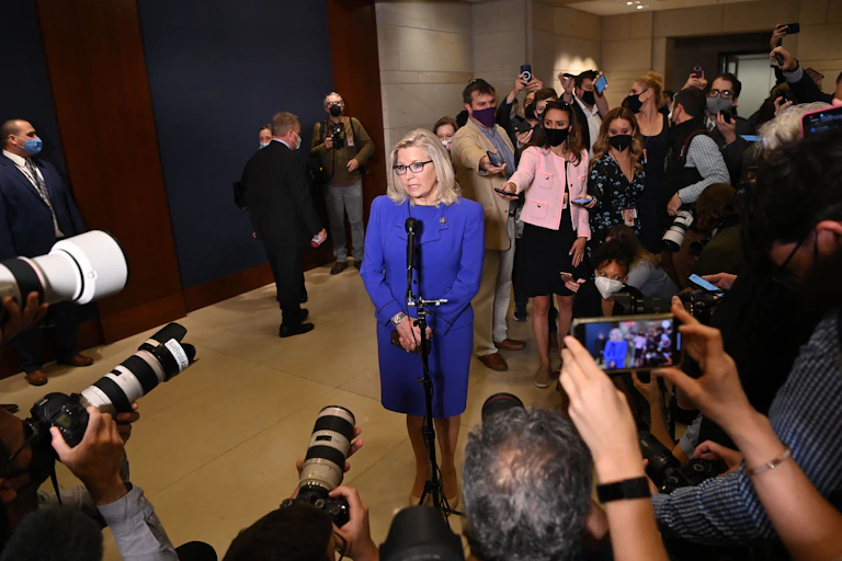 Rep. Liz Cheney, in a bright blue suit, speaking at a microphone while encircled by reporters and photographers.