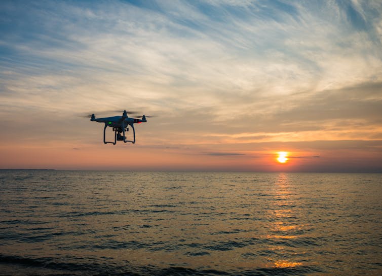 A drone is silhouetted against a sunset, with the sea beneath