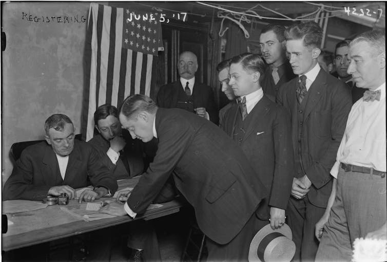 A group of men stand in line to fill out paperwork at a table staffed by other men
