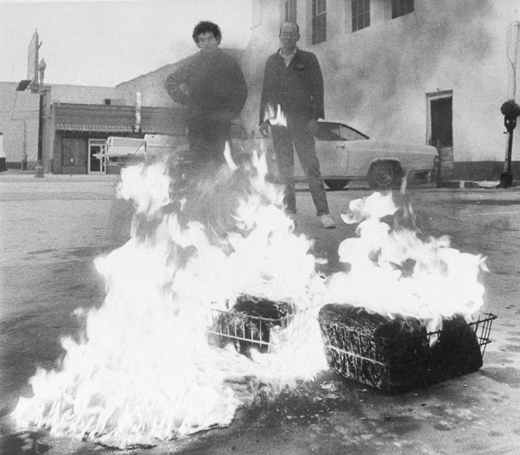 Dos hombres se paran frente a cajas en llamas.