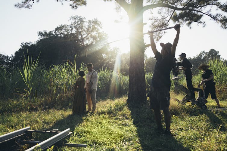 A set worker holds a boom in a sunny field while two actors act out a scene.