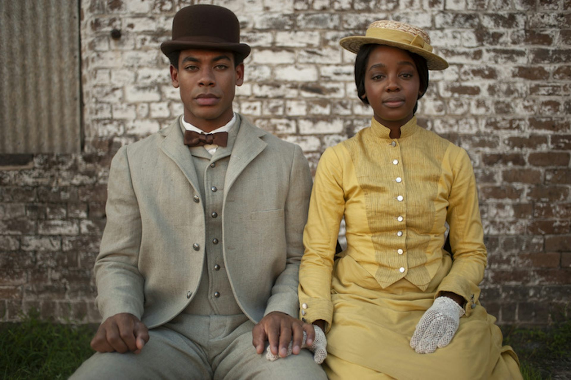 A man dressed in a dapper suit and hat poses, seated, next to a woman dressed in yellow.