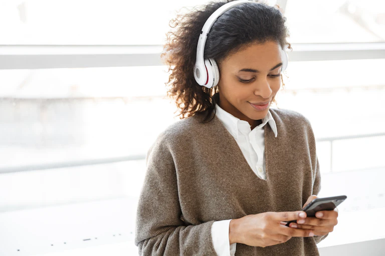 Une jeune femme devant une fenêtre écoute un balado sur son téléphone.