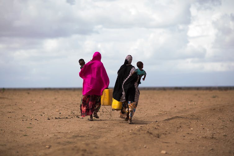 Two women carry small children and water jugs across a dry, empty landscape