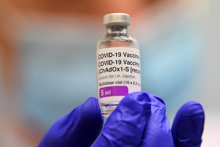 A gloved hand holds a vial of AstraZeneca vaccine.