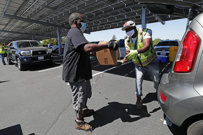Two men pass a box between them at a food bank next to a queue of cars.