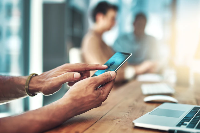 A man's hands holding a cell phone with a laptop in front of him.