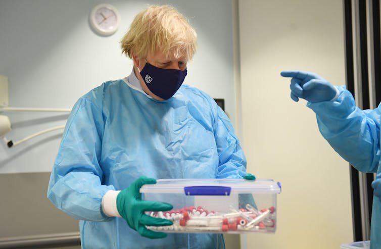 Boris Johnson wearing scrubs holding a box of PCR tests.