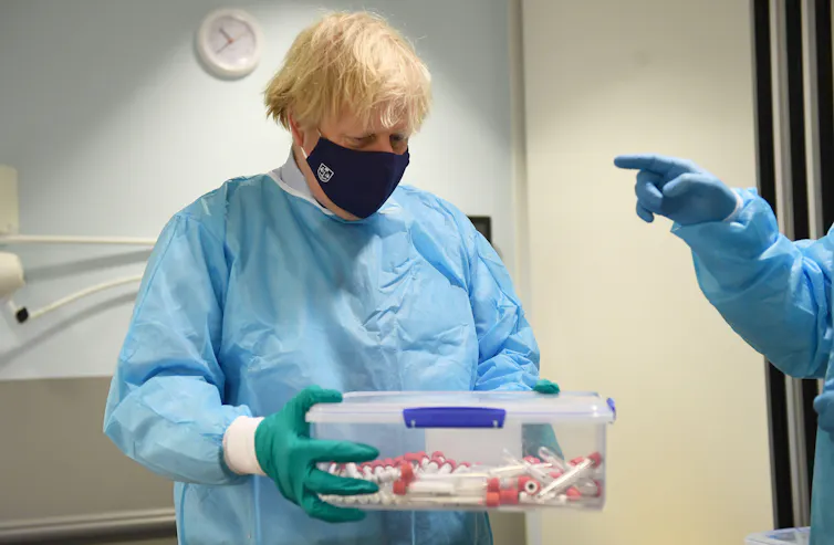 Boris Johnson wearing scrubs holding a box of PCR tests.