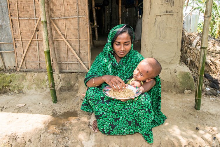 Femme assise à l'extérieur d'une cabane, tenant un petit enfant.