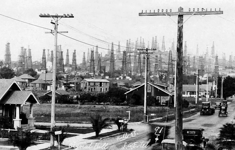 Une photo historique en noir et blanc montrant une rue avec des maisons, de vieilles voitures et des dizaines de derricks pétroliers sur la colline derrière elles