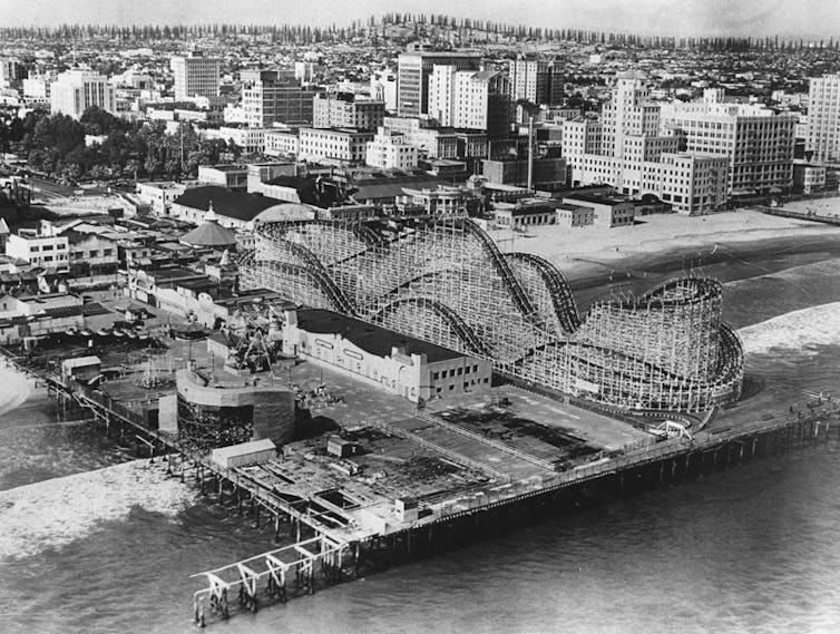 An old black-and-white photo of a roller coaster on a pier, with the city behind it and then a long row of oil derricks behind that on a ridge.