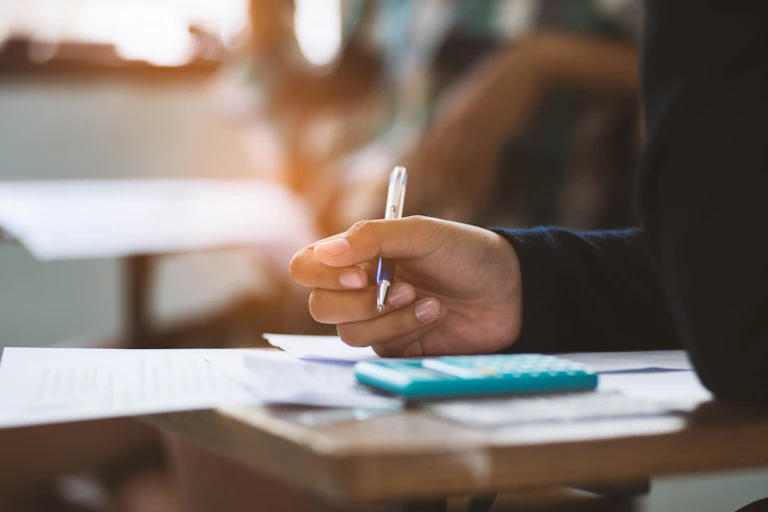 Student's hand writing a maths exam with calculator.