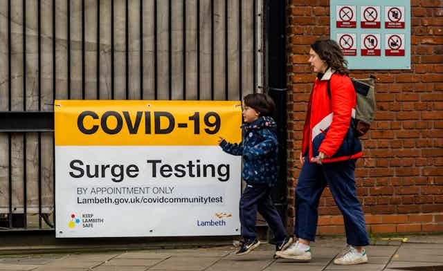 Mother and child walking past a COVID-19 Surge Testing sign in Oval, London.