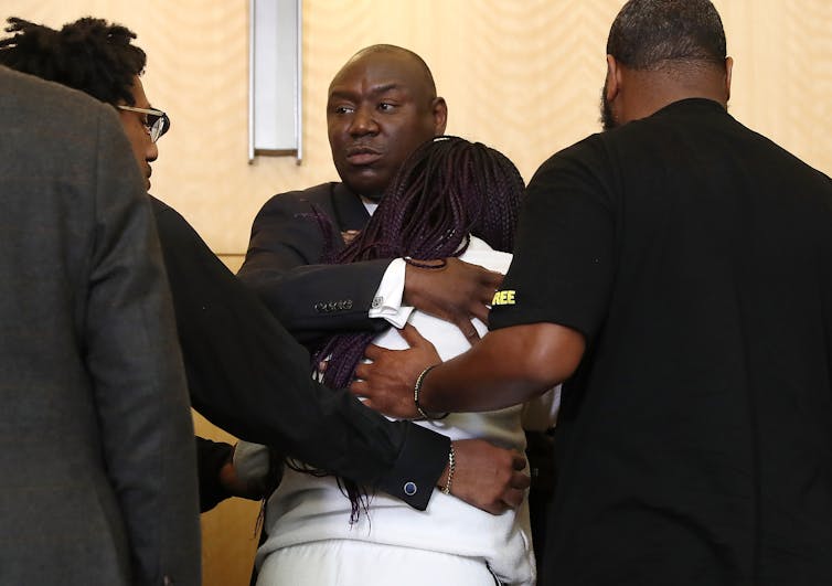 Black man facing the camera hugs a woman, whose back is to the camera, as other people physically support her