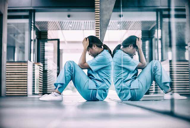 a nurse in scrubs sits on the floor with their head in their hands