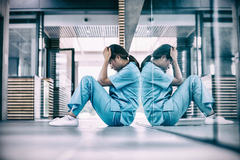 a nurse in scrubs sits on the floor with their head in their hands