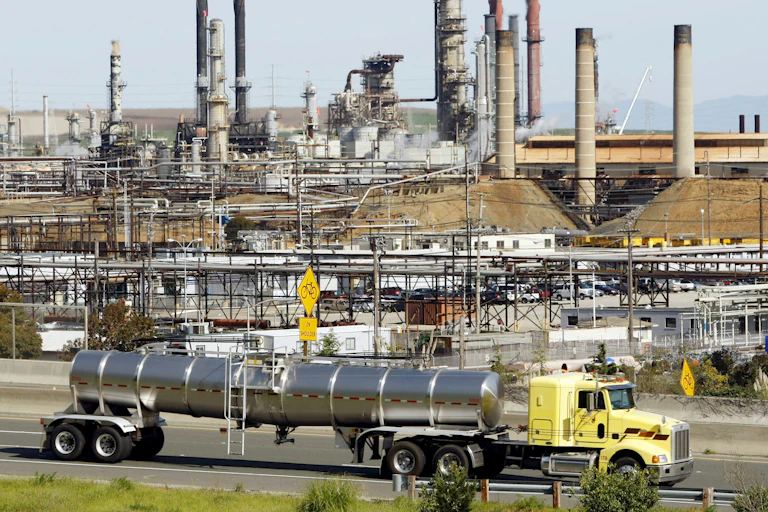 A tanker truck drives past a large refinery.