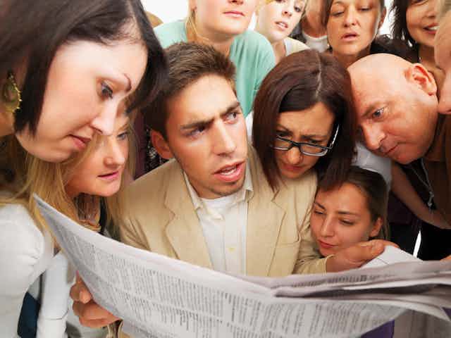A man with an intense look on his face is holding a newspaper with a lot of people around him looking at it.