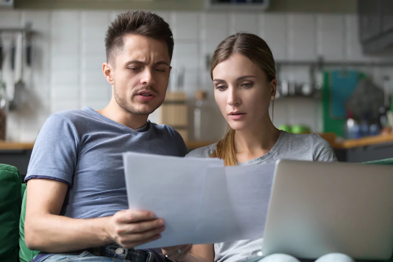 Couple in kitchen look at computer and papers.
