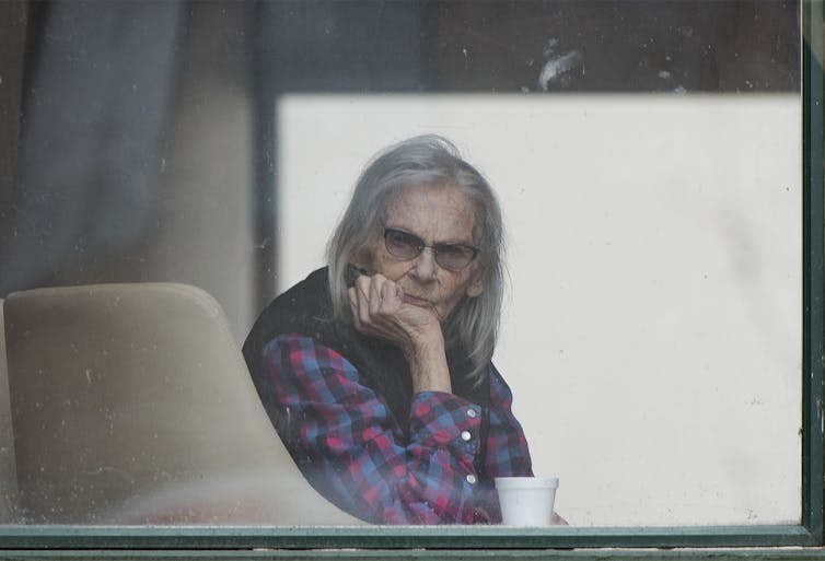 An elderly woman with long grey hair peers out the window of a long-term care home.