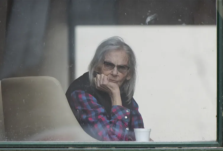 An elderly woman with long grey hair peers out the window of a long-term care home.
