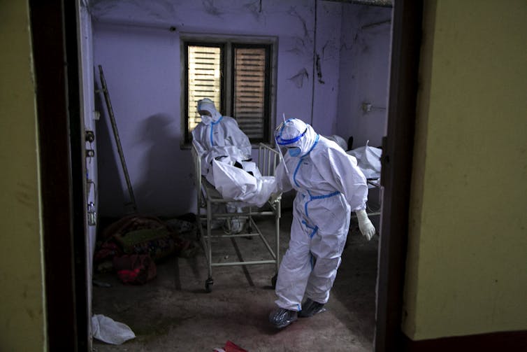 Paramedics in white hazmat suits roll a hospital gurney with a body covered in white sheet.