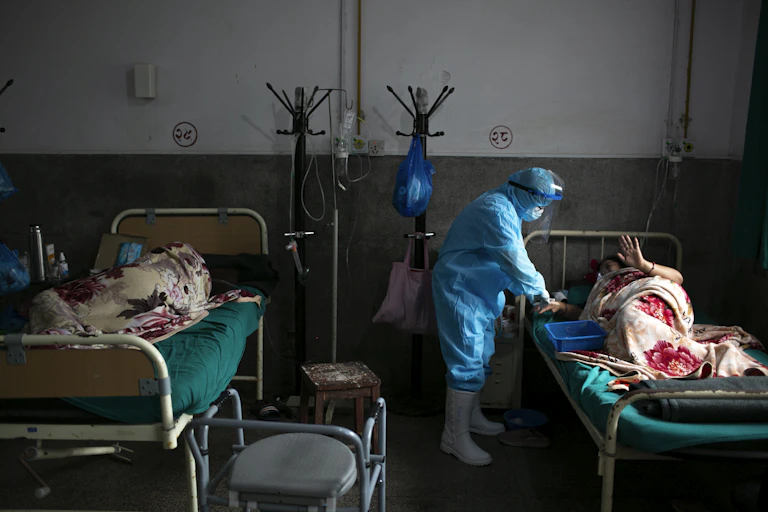 A nurse tends to a patient. The room shows two hospital beds, both occupied.