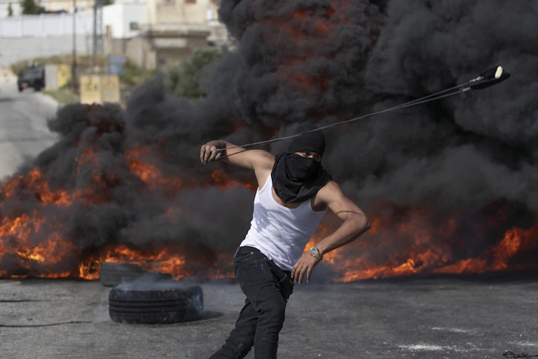A man in balaclava prepares to hurl a rock from a slingshot as tire fires burn in the background.
