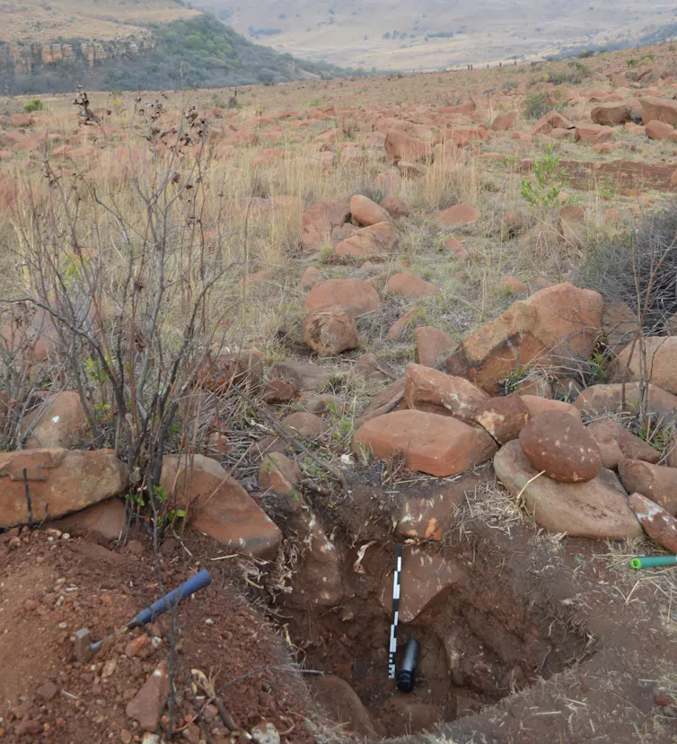 Some digging equipment and measuring sticks lie on the sandy ground while rolling mountains are pictured in the background