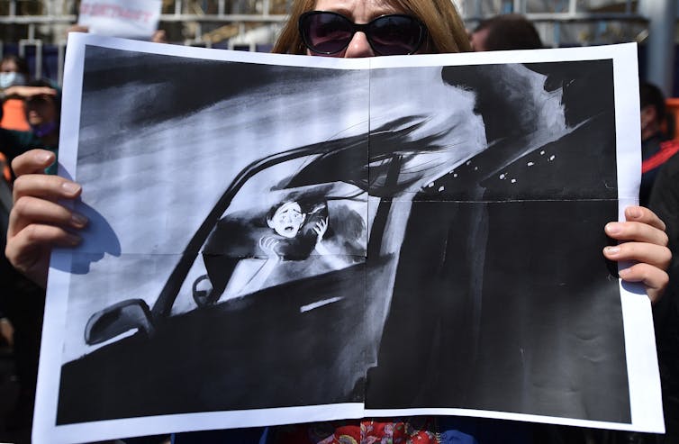 Woman holds a drawing depicting a scared woman being taken away in a car
