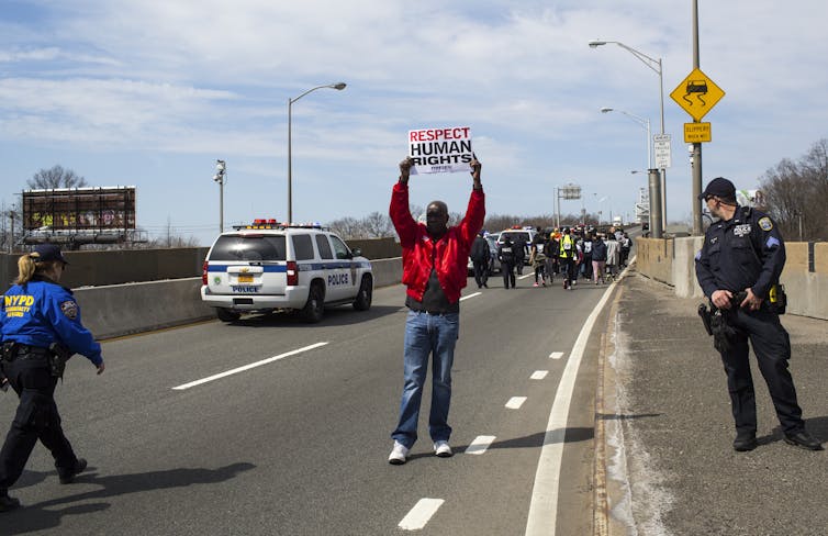 A man holding up a sign 'RESPECT HUMAN RIGHTS' as two police officers stand near him and approaching marchers.