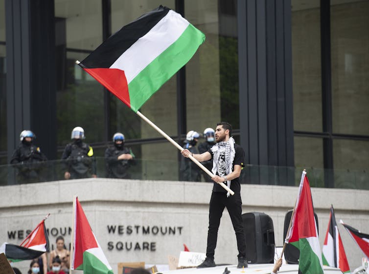 A man wearing a Keffiyeh around his neck stands above a crowd waving a Palestinian flag. Police officers stand in the background.