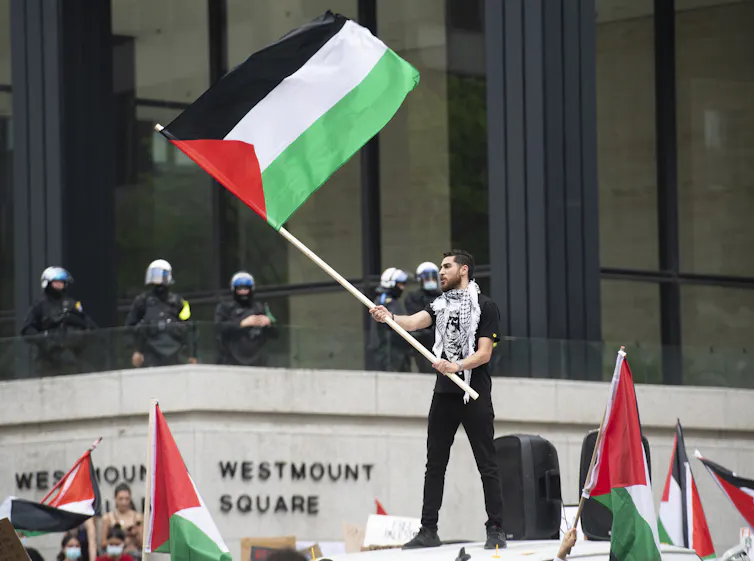 A man wearing a Keffiyeh around his neck stands above a crowd waving a Palestinian flag. Police officers stand in the background.