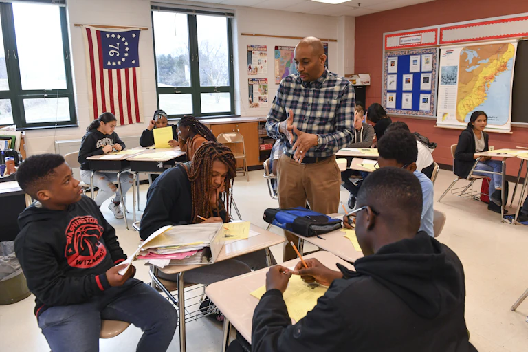 A teacher talks to a group of students sitting at desks.