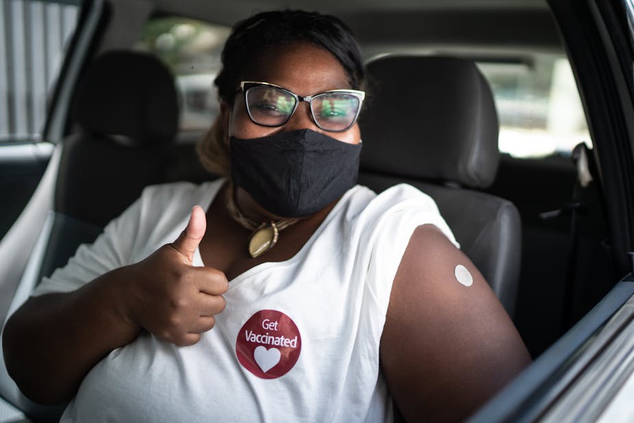 Person wearing face mask with a "Get Vaccinated" sticker on shirt, giving a thumbs up.