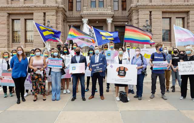 Protesters in front of a government building holding signs and flags supporting transgender rights. 