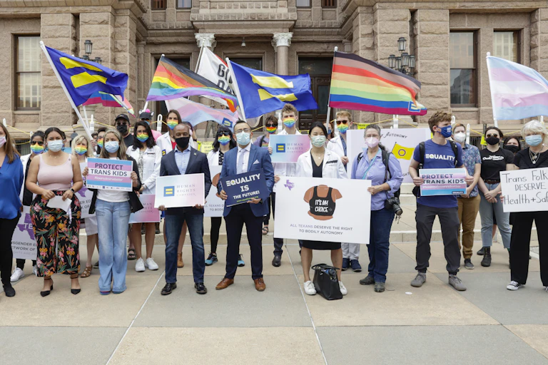 Protesters in front of a government building holding signs and flags supporting transgender rights.