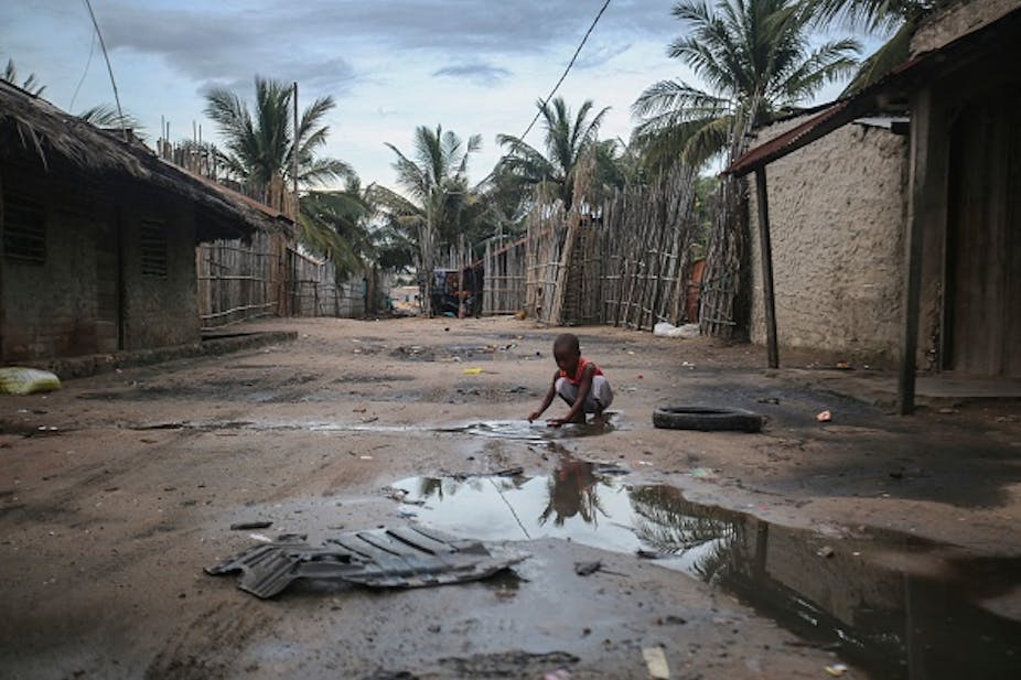 Child crouches on a muddy, puddled road between buildings, with palm trees in the background