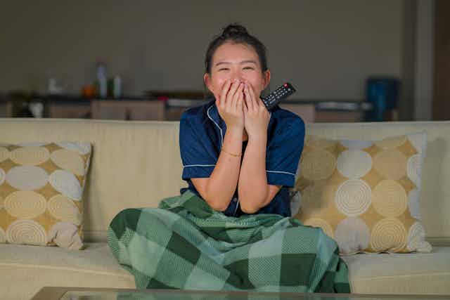 Woman laughing and covering mouth with remote in her hand while sitting cross legged on yellow sofa with green blanket on her lap