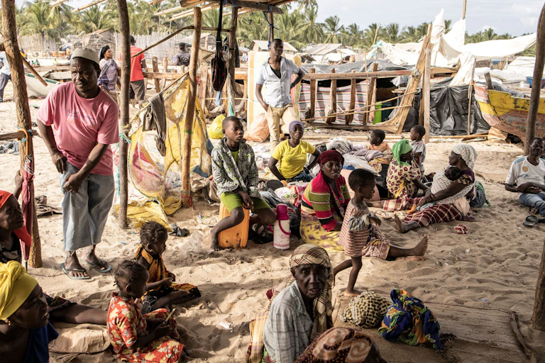 Men, women and children sit on beach sand in a makeshift shelter.