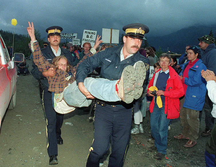 Two police carry a man by his legs and under his arms as a crowd looks on.