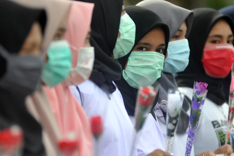 Mahasiswi memegang bunga mawar dan menggunakan masker sambil memperlihatkan spanduk kampanye Anti Kekerasan Seksual Terhadap Perempuan.