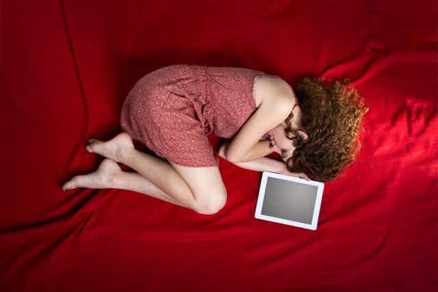 A woman in a red dress is curled up on a red sheet, a tablet beside her