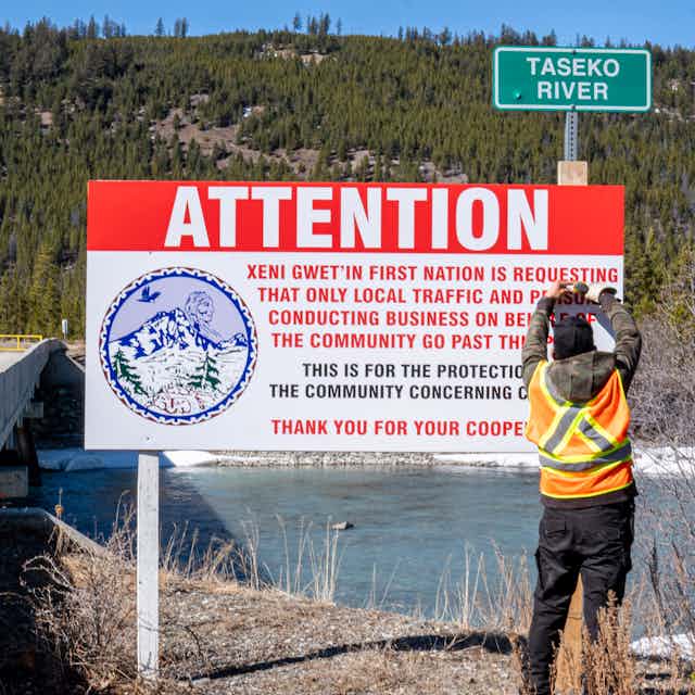 Man stands next to sign that reads 'Attention: Xeni Gwet'in First Nation is requesting that only local traffic and persons conducting business on behalf of the community go past this point'